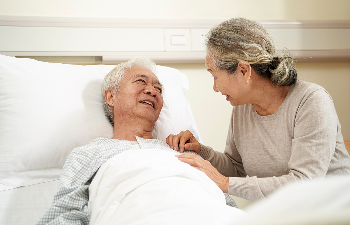 an elderly East Asian couple holding hands where one is in a hospital bed
