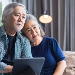"senior couple sit close on sofa, thinking, laptop"