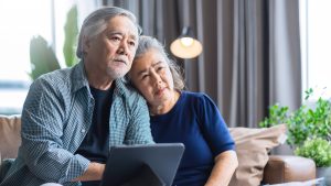 "senior couple sit close on sofa, thinking, laptop"