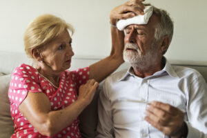 "A senior couple sitting on a couch. The wife is holding a cool, wet cloth to her husband's forehead to cool him off." 