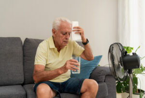 "Senior man sweating and holding glass water while sitting on sofa in front of electric fan at home."